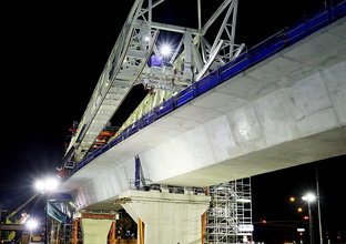 An on the ground view looking up at the construction work of the final span being lifted into place by a gantry crane at Sydney Metro's  Rouse Hill Drive site. 