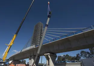 An on the ground view looking up at the construction of the railway bridge as cable poles are being lifted into place by a crane with the bridge tower in the background.