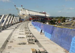 An on the ground view looking across at the construction of the railway bridge deck surface with rows of concrete slabs lined up to the right, pillars of concrete to the left and the large white gantry crane in the background.