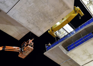 An on the ground shot looking up as the final segment of the Samantha Riley Drive bridge is being lifted into position with two construction workers guiding it in on a crane lift. 