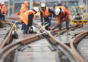 A group of four construction workers using machinery to lay the train tracks at a cross section, with more constructions completing the same task in the background. 