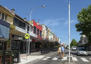 An on the ground shot stood in the centre of a two-way road looking down Marrickville high street with two people crossing at the pedestrian crossing with a church in the distant background. 