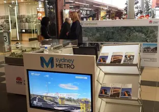 A close up view of the Sydney Metro mobile community stand showing a TV with display on it, brochures, train model and alignment map as community members are looking at the display behind. 