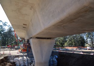 A close up view under the bridge as construction work continues on Sydney Metro's skytrain platform, there are three heavy machinery in the background behind a fence of the platform construction. 