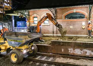 Two construction workers in orange high-vis watch an excavator digging while performing track works at Canterbury Station.