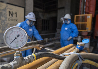 A close up view of a pressure gauge while two men in full PPE suits are in the background.