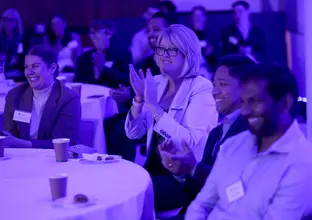 Audience is sitting at tables smiling and clapping while listening to a presentation.