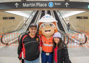 Couple with the Metro Mascot in front of the escalators on the platform