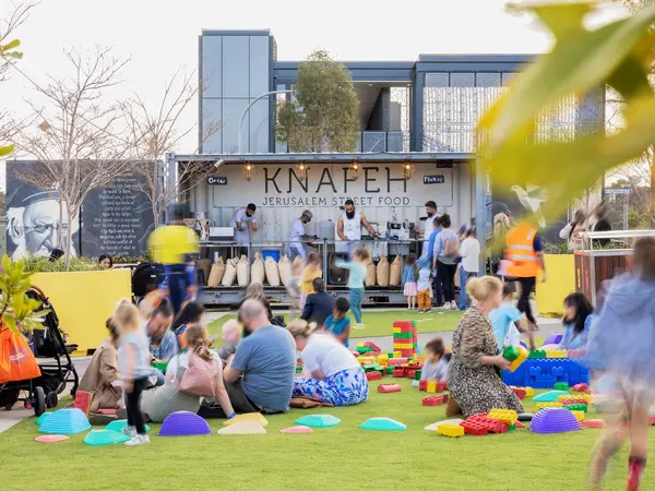 Families sitting down in a park playing with colourful blocks. A Jerusalem street food stand is in the background.