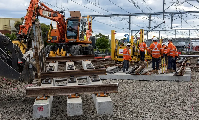 An on the ground view looking at a section of train tracks laid on top of a concrete slab that will be lifted into place by the crane digger with a group of five construction workers in the background working on the track line behind between Sydney Metro's Sydenham and Bankstown Stations. 