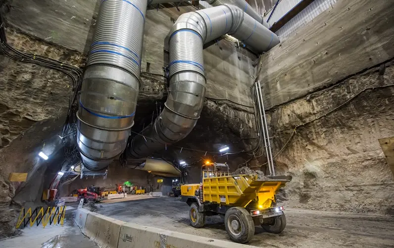 An on the ground view looking across to the tunnel at Sydney Metro's Pitt Street North construction site with two large pipes running through and out of the tunnel and a Roadheader about to drive into the tunnel. 