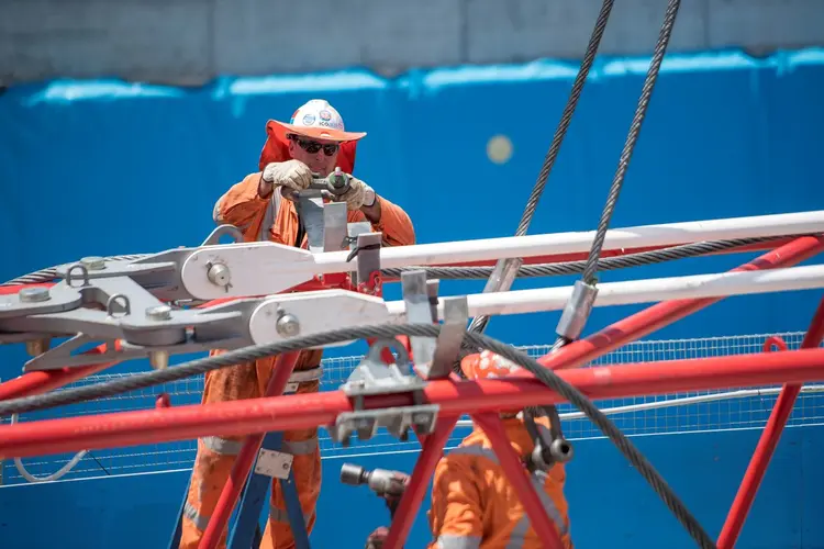 Two construction workers in high viz working on a large crane. The wall of the station box is in the background.