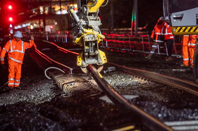 The arm of a heavy machine is picking up and moving the rail track at night time. Construction worker in high viz is directing the operator of the machine. 