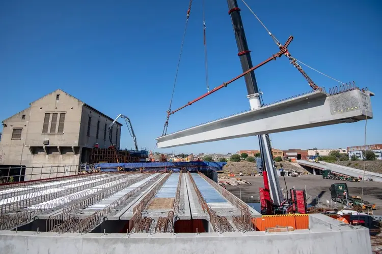 A crane lifting a beam into place at the Sydenham Station and Junction site. There is a building and blue sky in the background.