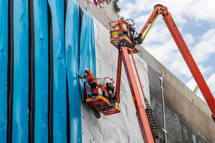 Workers in high-vis are on two cherry picker lifts installing blue waterproof sheeting to the walls of Waterloo Station box.