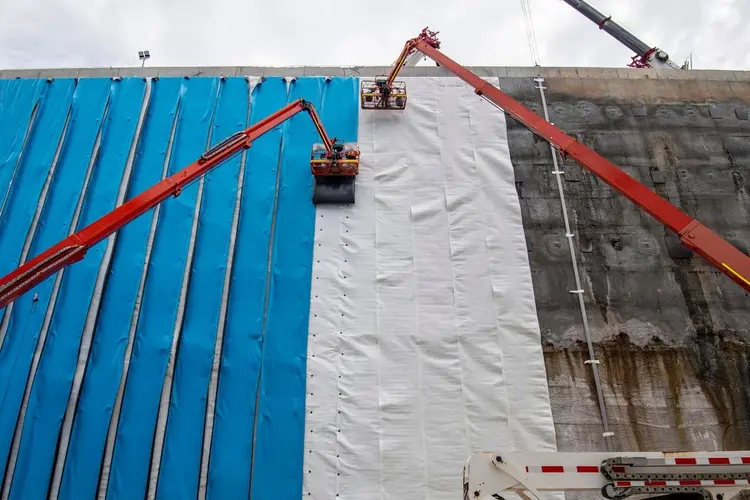 Workers in High Viz are on two Cherry picker lifts installing blue waterproof sheeting to the walls of Waterloo Station box.