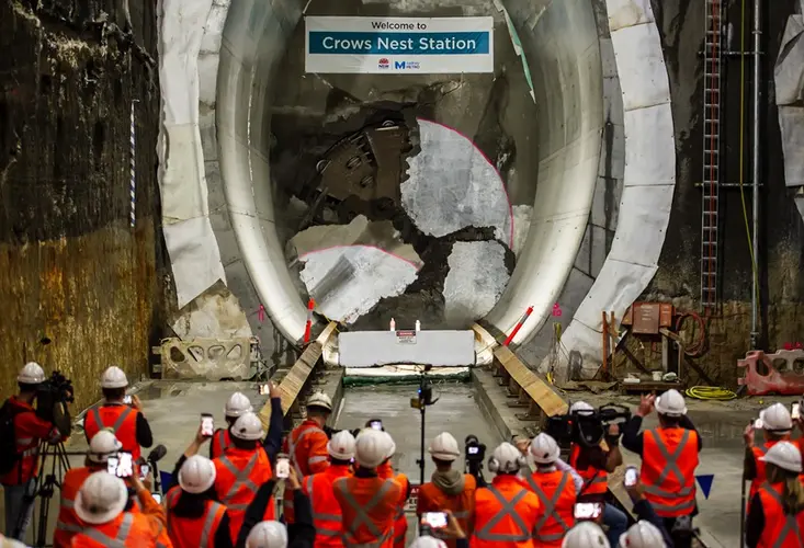 An on the ground shot behind a large group of construction workers watching as Tunnel Boring Machine breaks through the final section of the wall at Sydney Metro's Crows Nest Station. 