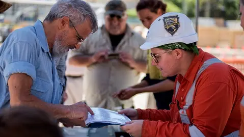 Sydney Metro employees showing community members the archaeological artefacts found at Waterloo Station as they both point to a booklet labelling the displayed artefacts on the table below them. 