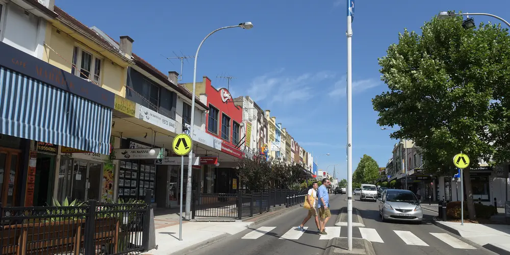 An on the ground shot stood in the centre of a two-way road looking down Marrickville high street with two people crossing at the pedestrian crossing with a church in the distant background. 