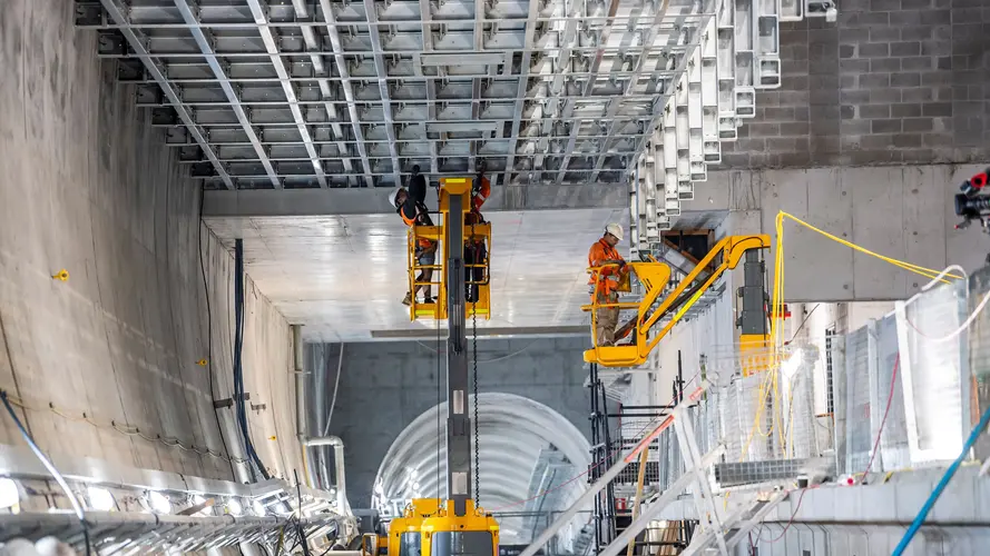 Two construction workers standing on a cherry picker lift while installing the ceiling inside Victoria Cross Station.