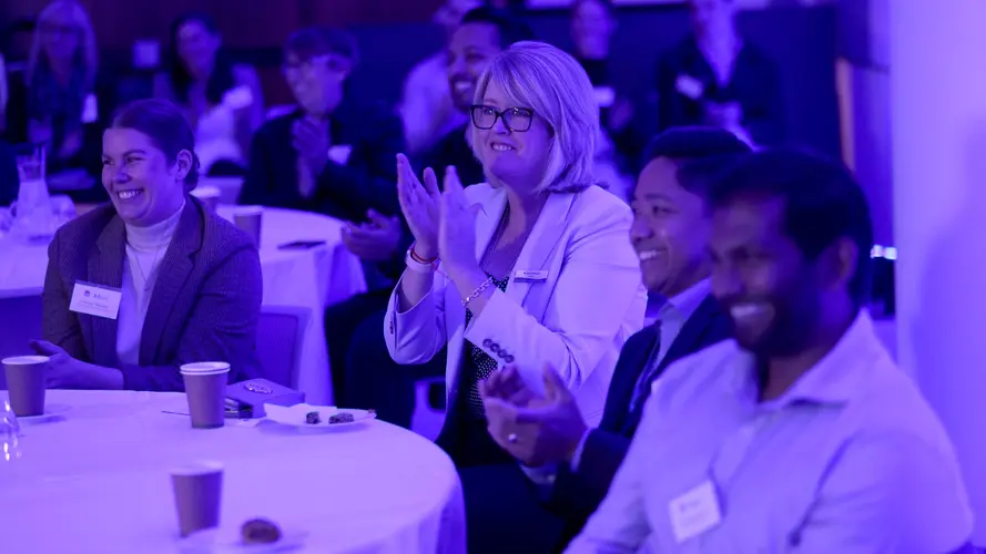 Audience is sitting at tables smiling and clapping while listening to a presentation.