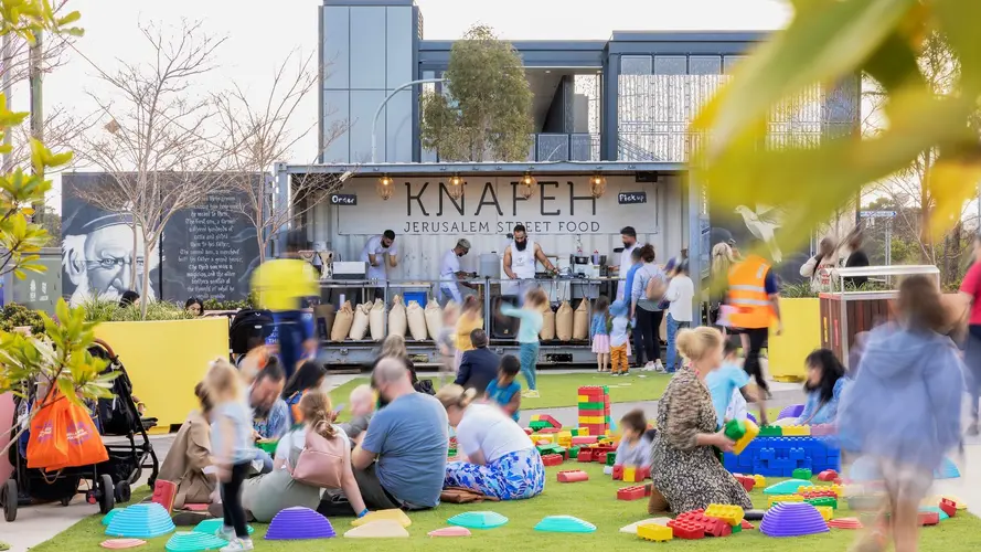 Families sitting down in a park playing with colourful blocks. A Jerusalem street food stand is in the background.