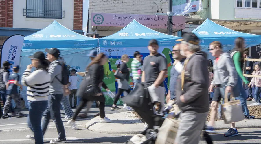 A group of community members walking past three Sydney Metro marquees at a community event